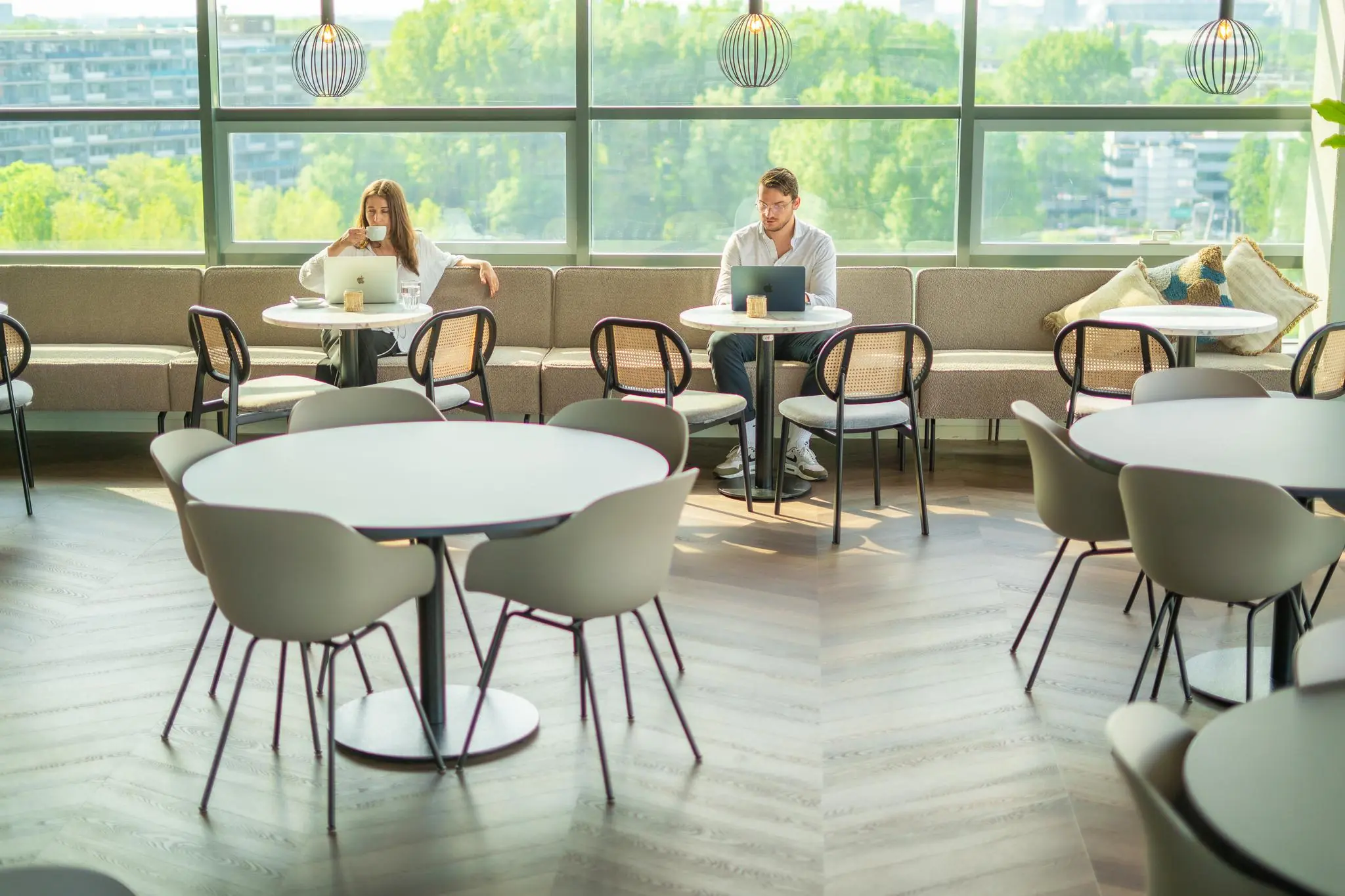 Twee mensen werken op laptops en drinken koffie in een moderne lounge aan de Joop Geesinkweg met uitzicht op groen.